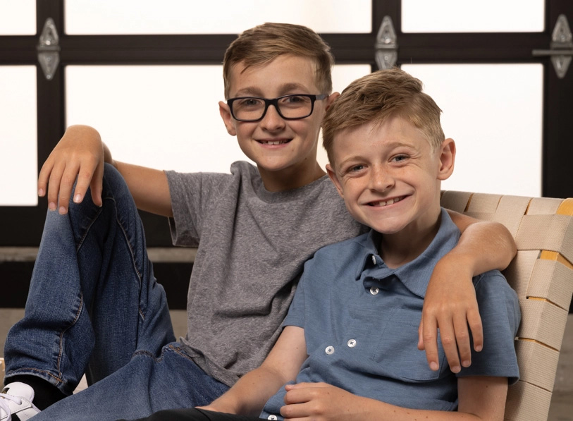 Two young boys sit together in a chair; one boy has an arm across the shoulders of the other boy.